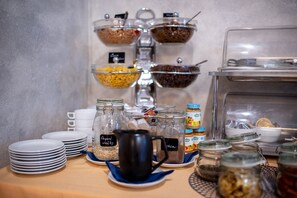 A buffet setup with various food items in glass containers, a coffee urn, and stacked plates. - Felicity Garni Hotel (Bratislava)