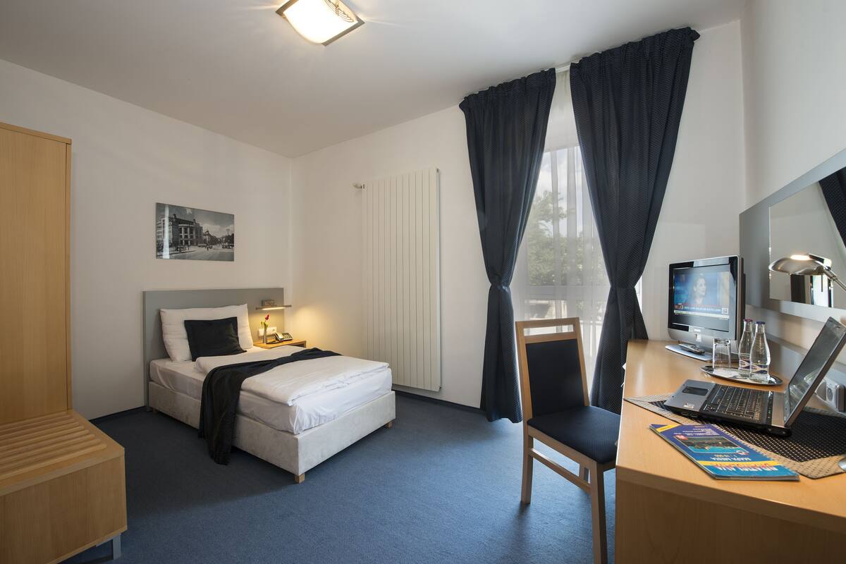 Una habitación de hotel con cama, escritorio, silla, computadora y ventana con cortinas.