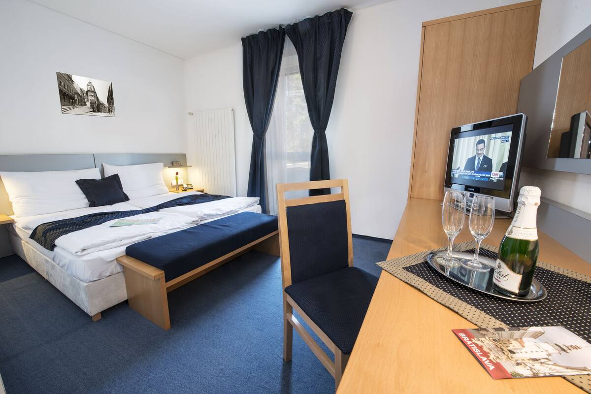 Habitación de hotel con una cama, una silla, una mesa con una botella y vasos, y un televisor mostrando un noticiero.