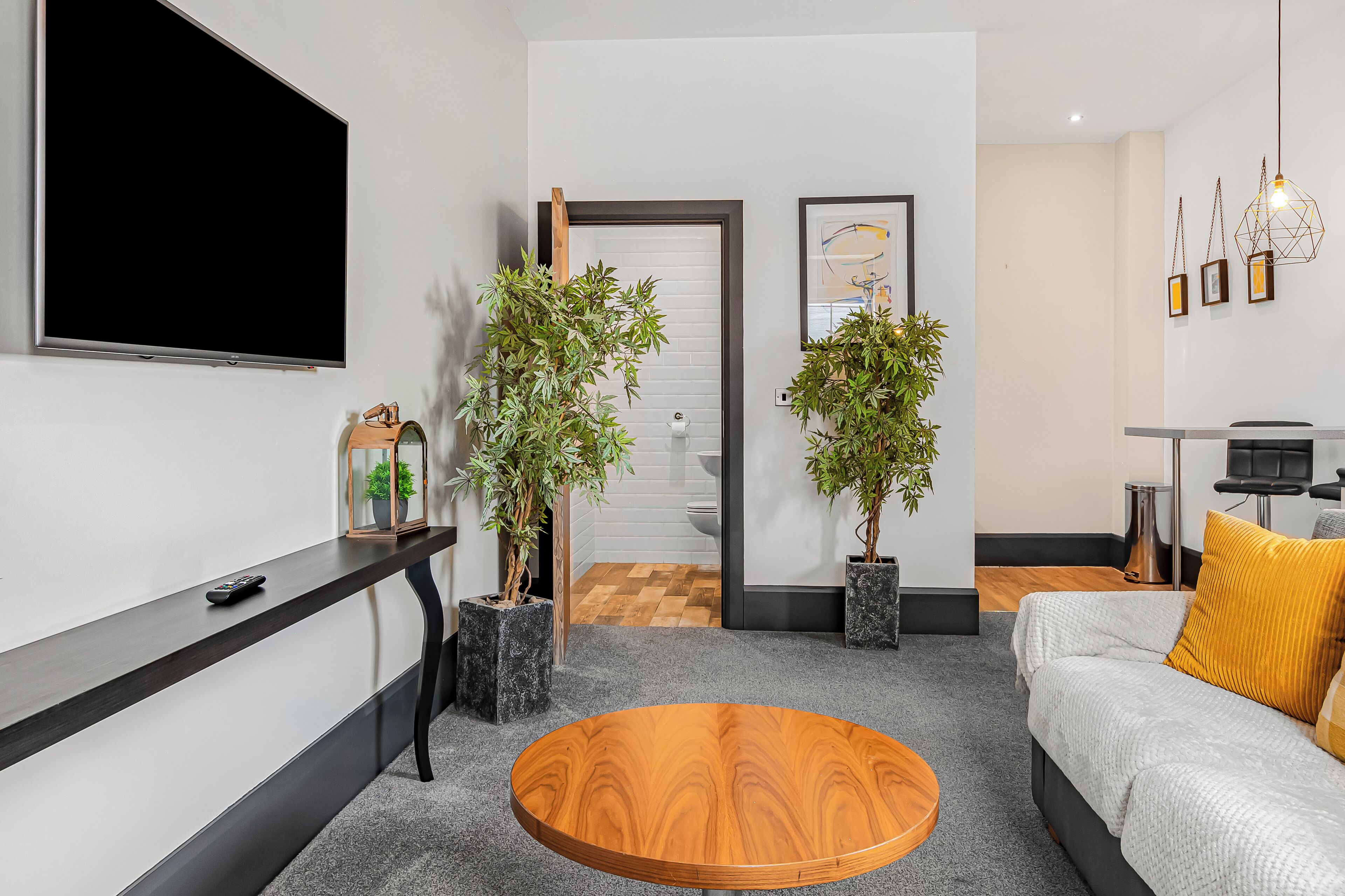A modern living room with a large flat-screen TV, a round wooden coffee table, a sofa with a yellow pillow, and two potted plants.