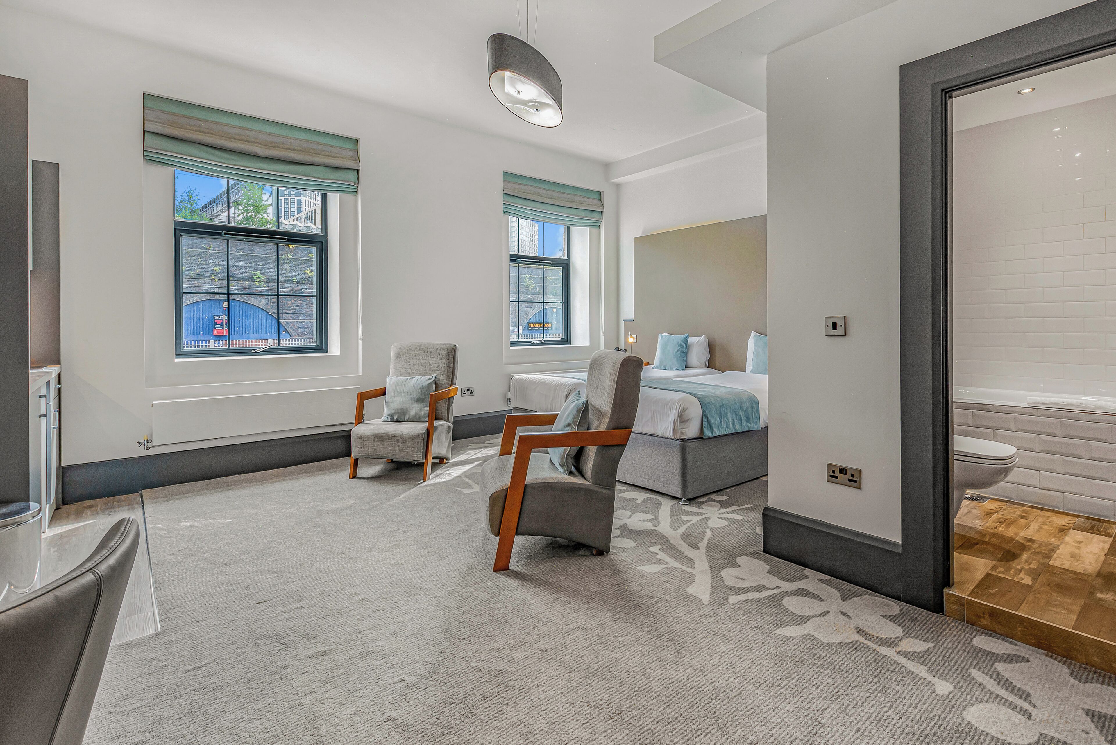 Ein modernes Hotelzimmer mit einem großen Bett, zwei Sesseln, einem Schreibtisch und Blick auf die Stadt durch große Fenster.