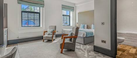 Ein modernes Hotelzimmer mit einem großen Bett, zwei Sesseln, einem Schreibtisch und Blick auf die Stadt durch große Fenster.