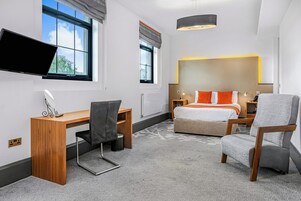Una habitación de hotel moderna con un escritorio de madera, una silla cómoda, una cama con almohadas naranjas, un ventanal grande y un televisor de pantalla plana.