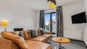 A modern hotel room with a leather sofa, a round table, a TV mounted on the wall, and a view of the cityscape through large windows. - The Ainscow Hotel (Salford)