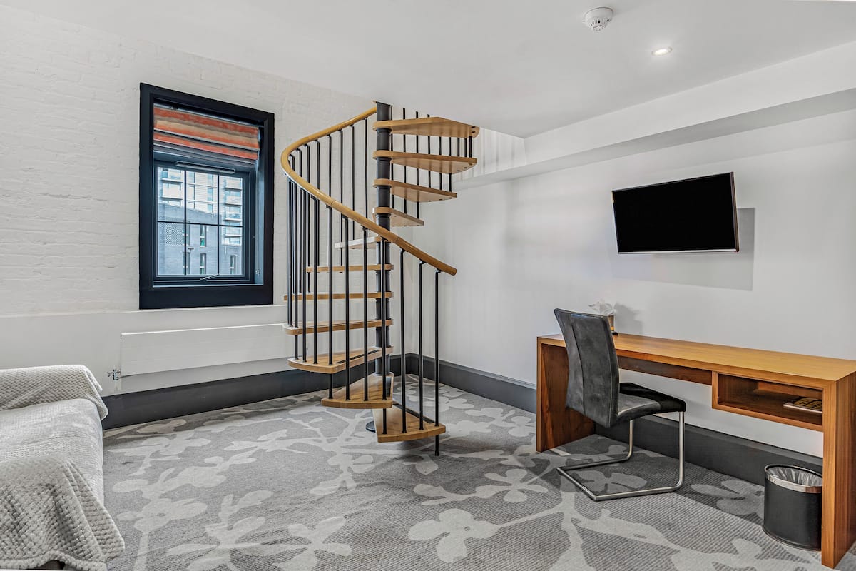 Una habitación moderna con una escalera espiral, un escritorio con una silla, un televisor y una ventana con persianas.