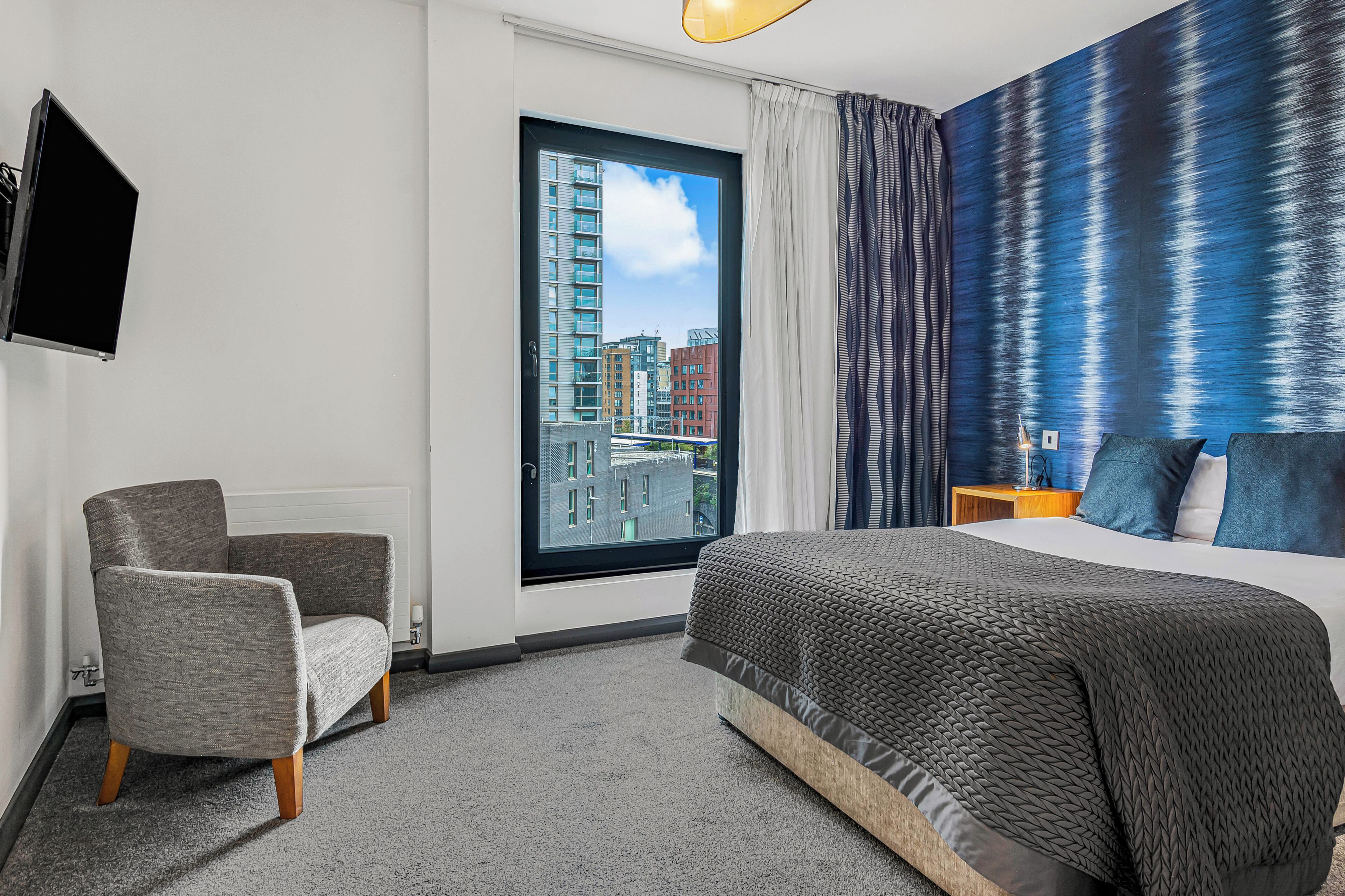 A modern hotel room with a large bed, a grey armchair, a flat-screen TV, and a city view through the window.