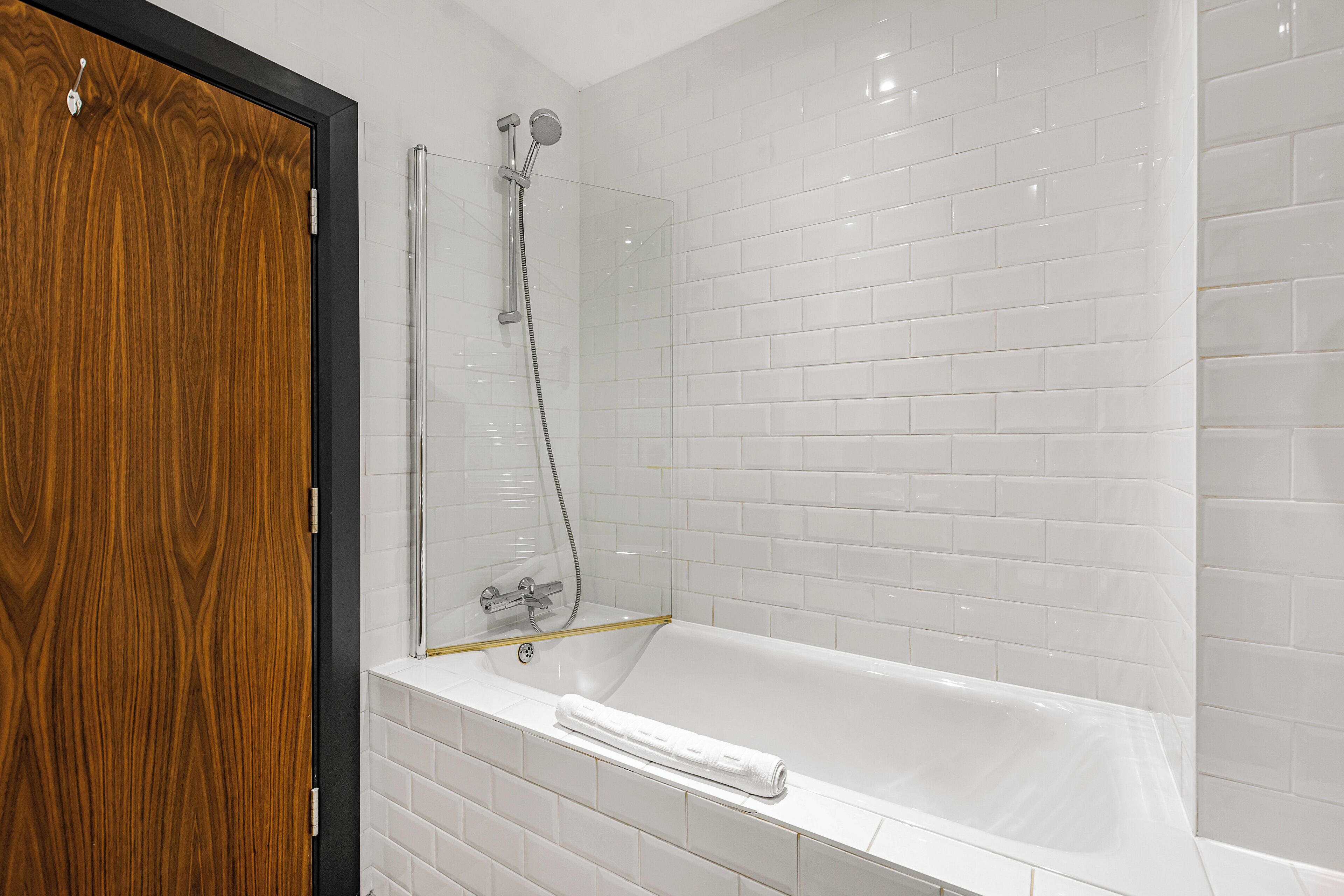 A modern bathroom with a white-tiled shower enclosure, a wooden door, and a white bathtub with a towel.