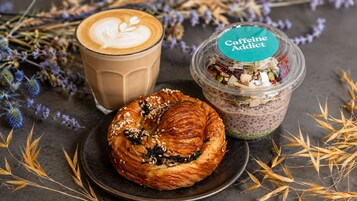 Un croissant con semillas, una taza de café y un recipiente con la etiqueta 'Caffeine Addict' junto a varios bocadillos.