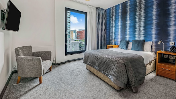 A modern hotel room with a large bed, a grey armchair, a view of the city through a large window, and a textured blue wall. - The Ainscow Hotel (Salford)