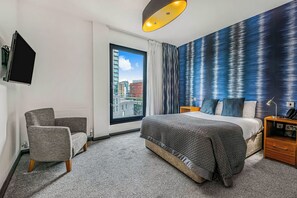 A modern hotel room with a large bed, a grey armchair, a view of the city through a large window, and a textured blue wall. - The Ainscow Hotel (Salford)