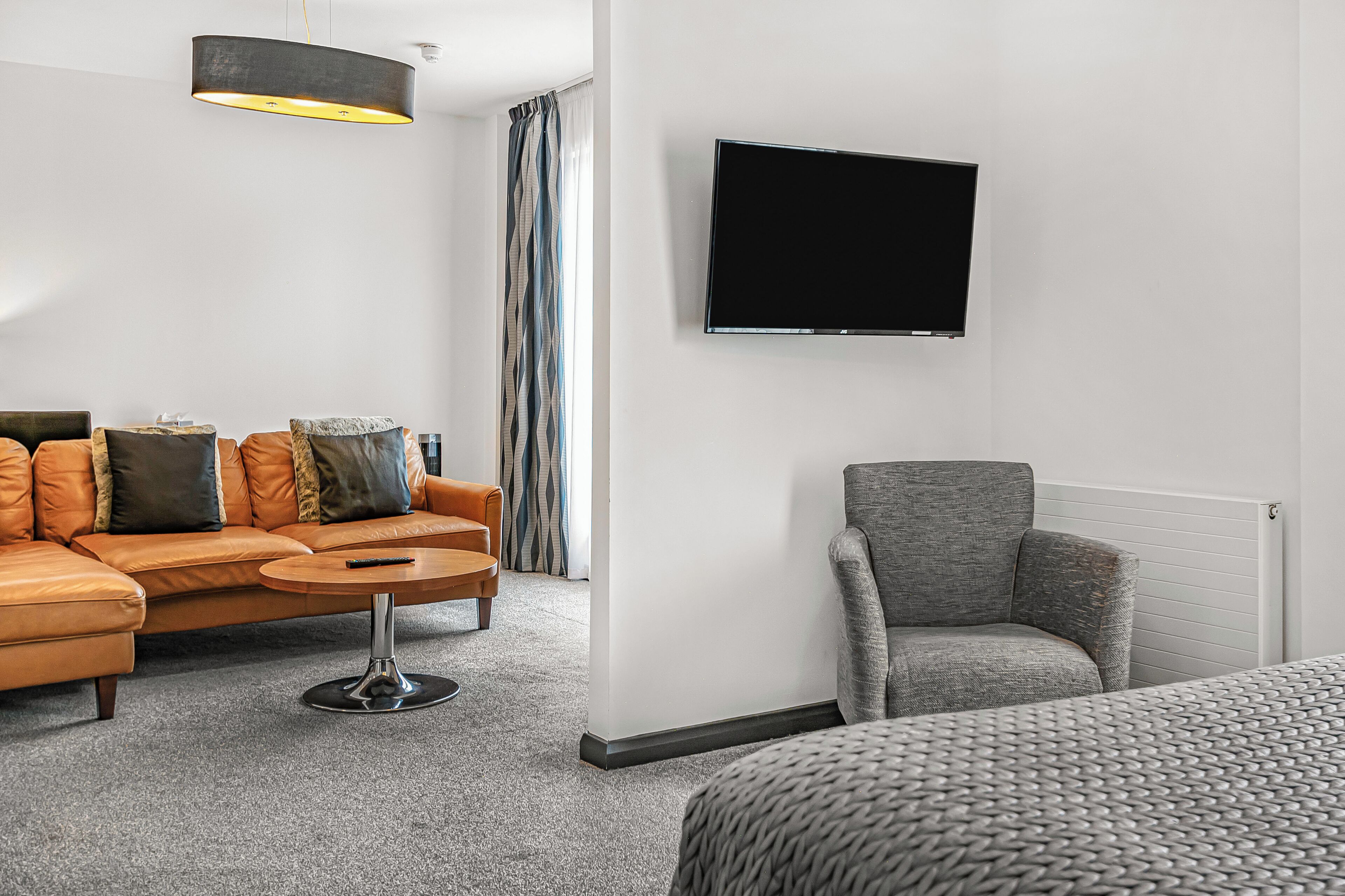 A modern hotel room with a sofa, armchair, and a flat-screen TV mounted on the wall.