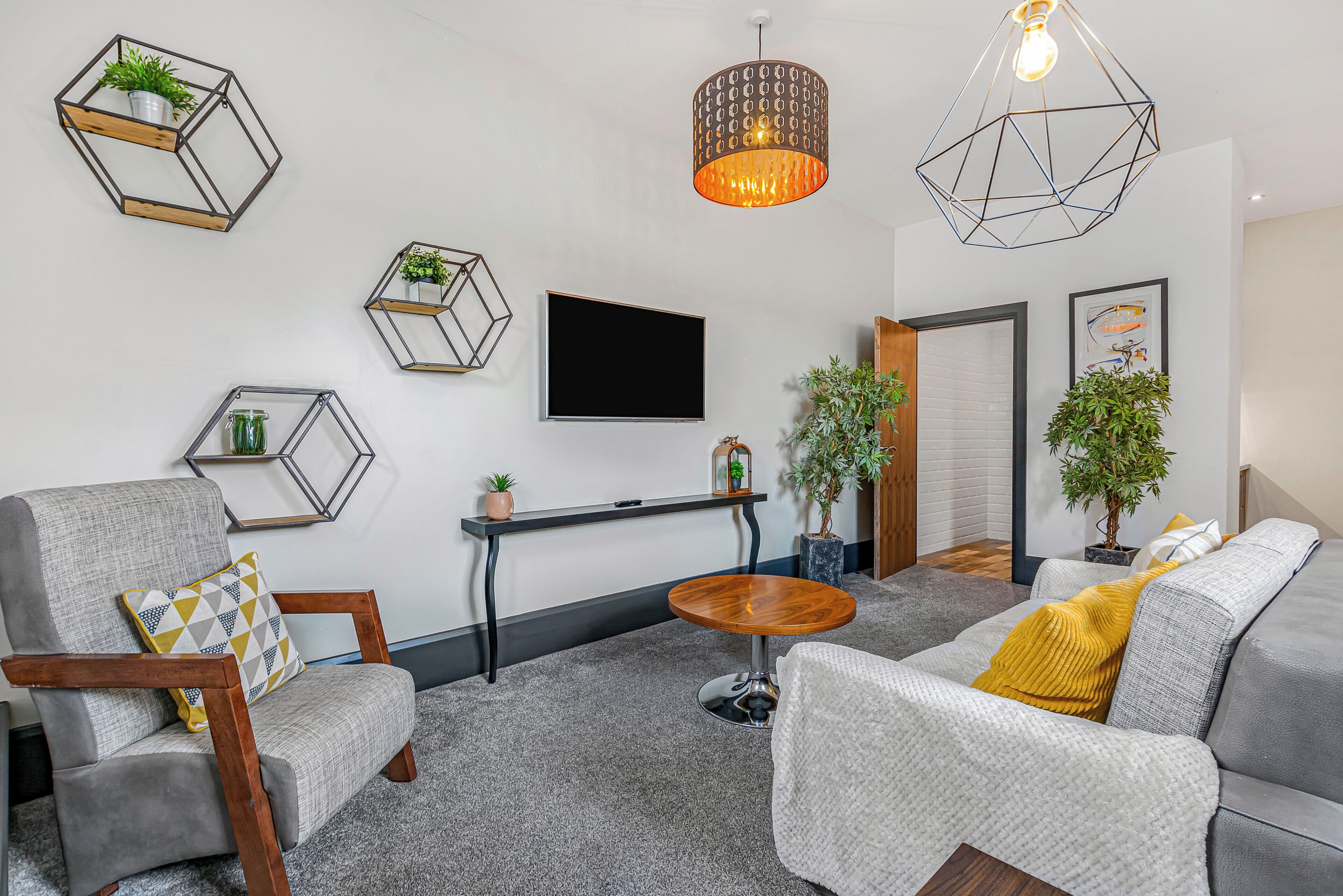 A modern living room with a grey sofa, a wooden chair, a small round table, and hexagonal wall shelves with plants.