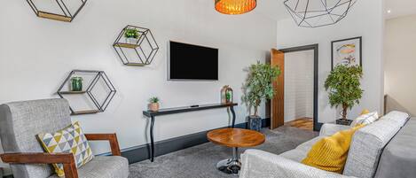 A modern living room with a grey sofa, a wooden chair, a small round table, and hexagonal wall shelves with plants.