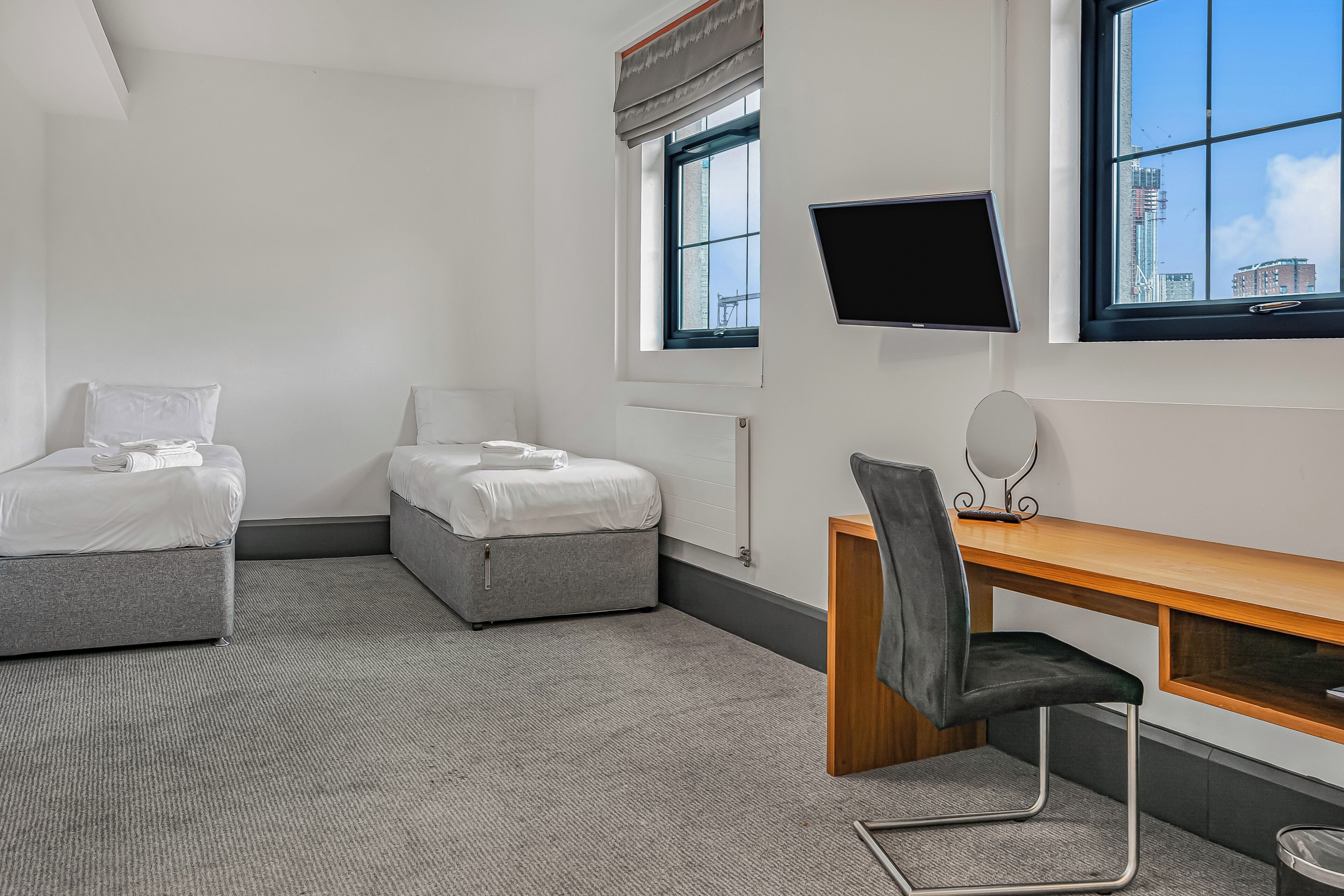 Ein Hotelzimmer mit zwei Betten, einem Schreibtisch, einem Stuhl, einem Fernseher und einem Fenster mit Blick auf die Stadt.