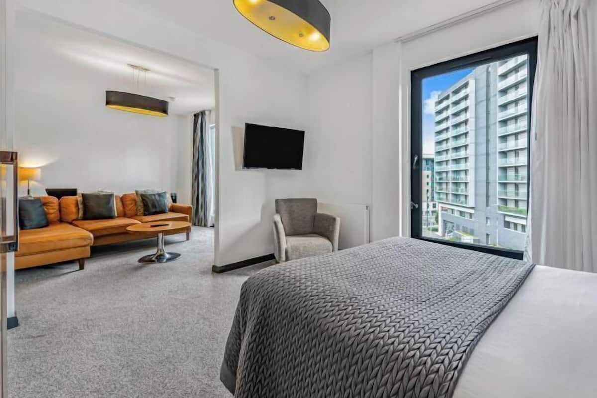 Habitación de hotel con cama, sofá, sillón, televisión y ventana con vista a la ciudad.