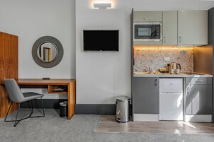Una cocina compacta con microondas, refrigerador y pileta. Hay un cubo de basura pequeño y un escritorio de madera con una silla.