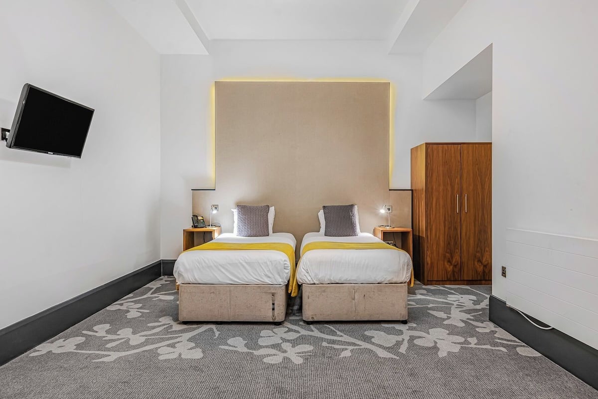 Habitación de hotel con dos camas, un armario de madera, un televisor de pantalla plana en la pared y una alfombra estampada.