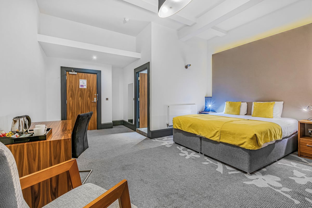 Una habitación de hotel con una cama, un escritorio con silla, una cafetera y una mesita de noche con lámpara.