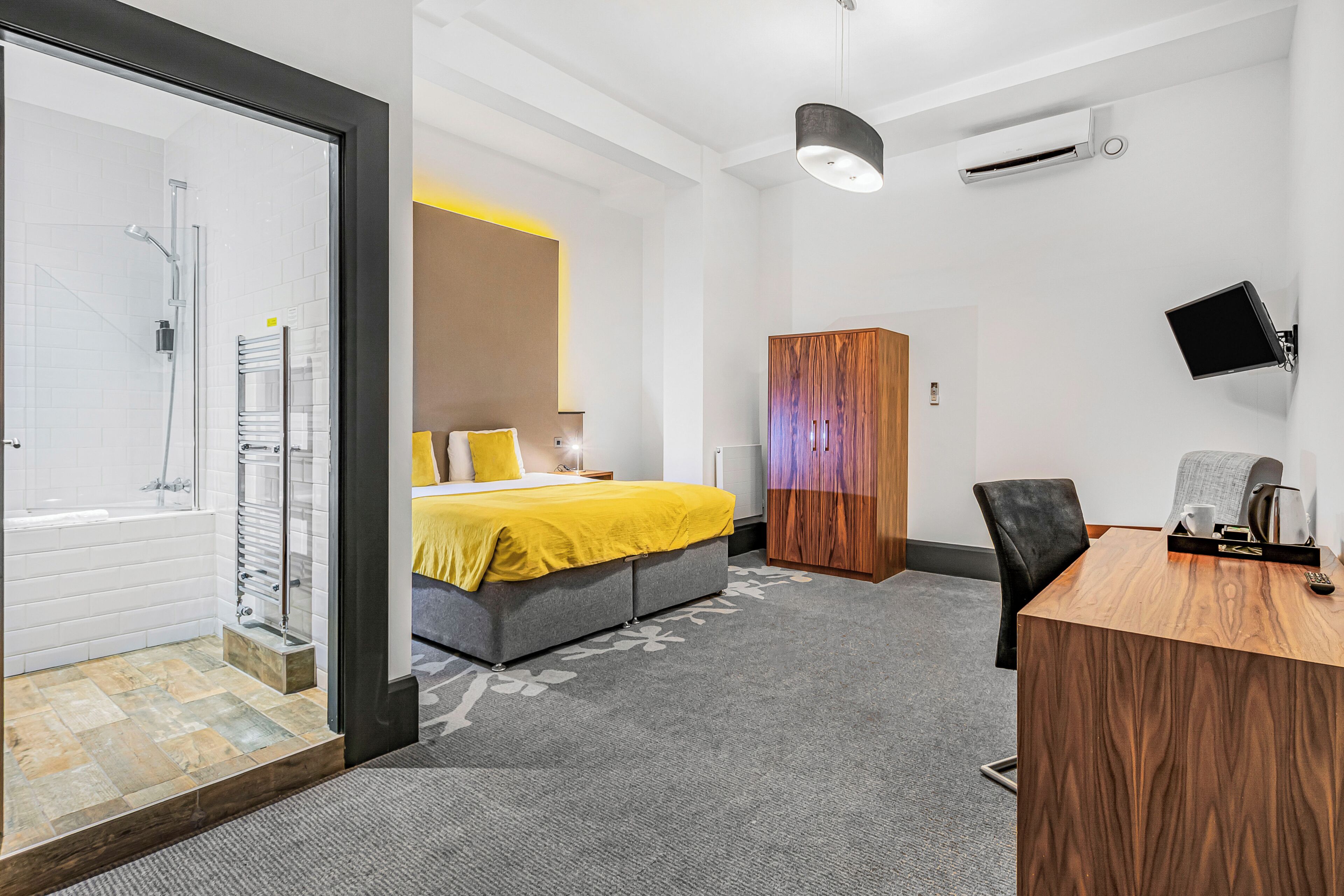 Habitació d'hotel moderna amb dutxa, un llit amb llençol groc, un armari de fusta, escriptori amb cadira i televisió penjada.
