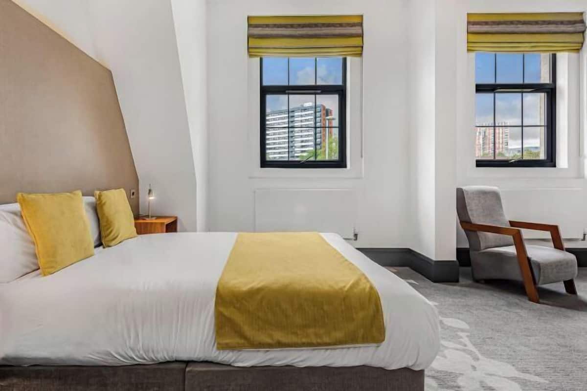 Habitación de hotel con cama, dos ventanas con persianas, una silla y vistas a edificios.