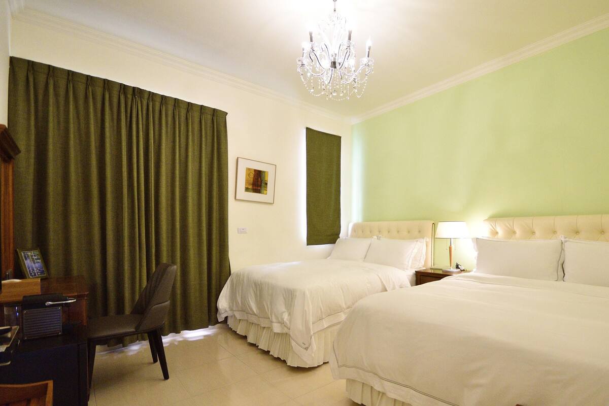 A hotel room with two beds, a desk, a chair, a chandelier, and a painting on the wall.