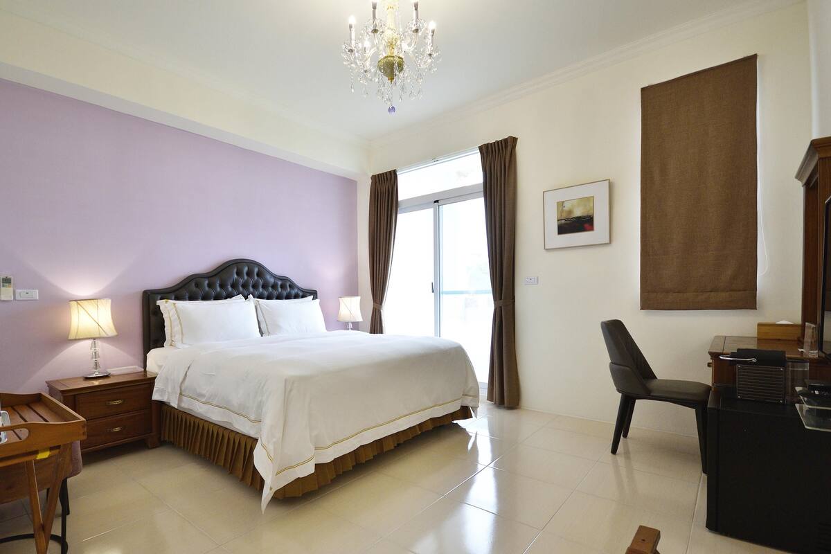 A hotel room with a bed, bedside table, lamp, desk, chair, and a large window with curtains.