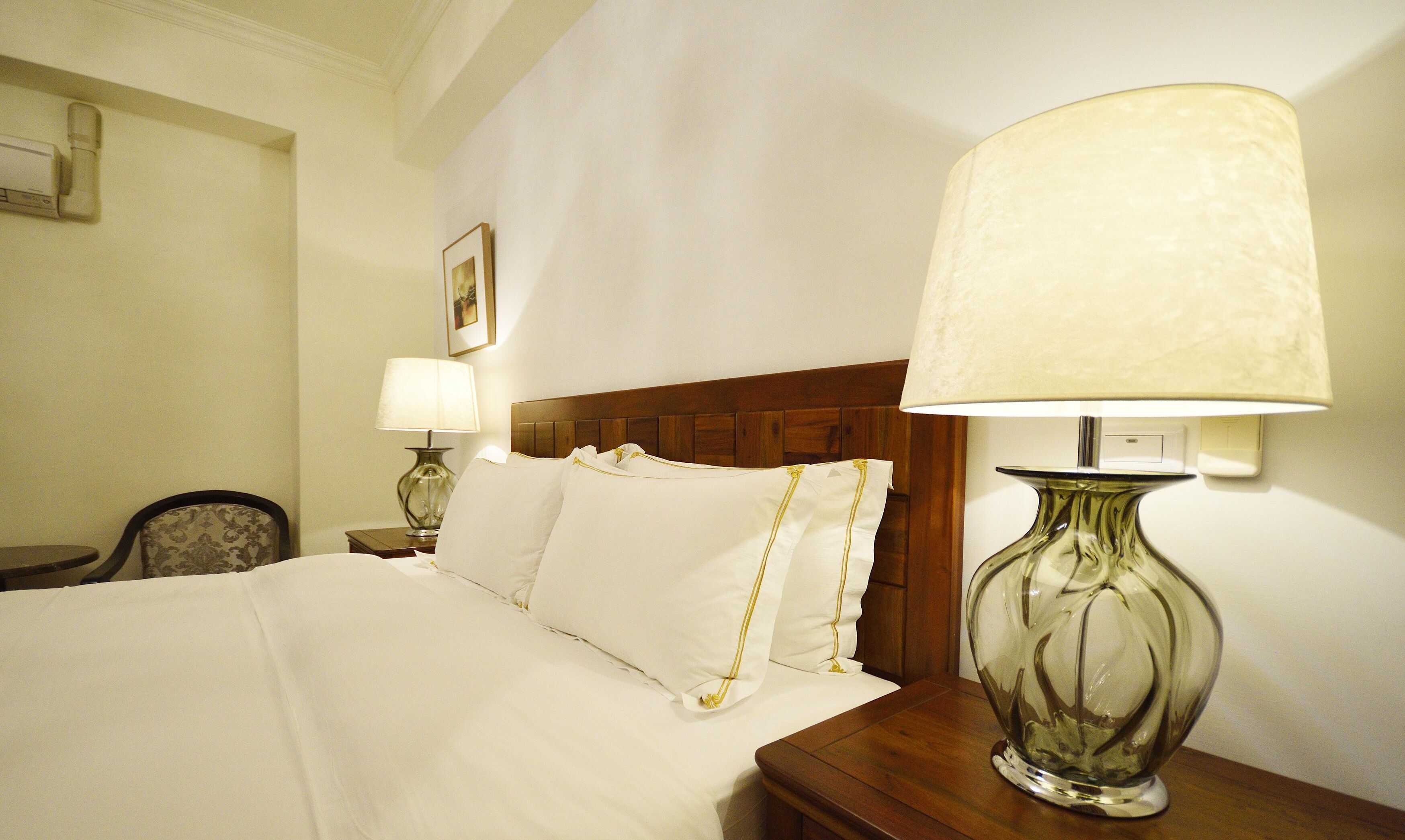 Una habitación de hotel con una cama bien hecha, un cabecero de madera, una mesita de noche con lámpara y una silla.