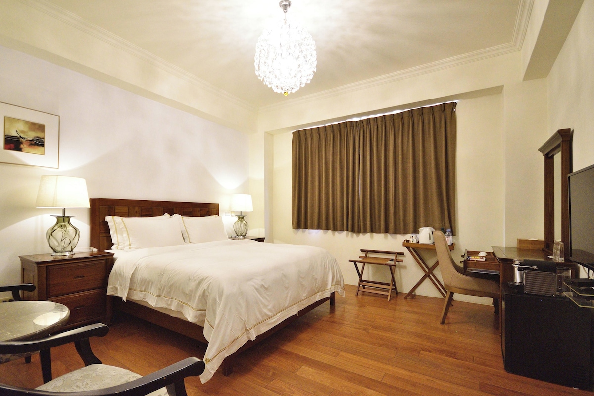 Una habitación de hotel con cama, mesitas de noche, escritorio, silla, televisión y ventana con cortinas.