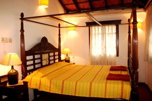 Uma cama com dossel, com colcha listrada e duas almofadas vermelhas, ladeada por mesas de cabeceira com abajures, em um quarto com teto de madeira e janela com cortinas leves.