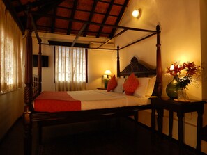 Uma cama com dossel e cabeceira de madeira, um cobertor vermelho e dois travesseiros vermelhos. Há um vaso com flores em uma mesa de cabeceira ao lado da cama.