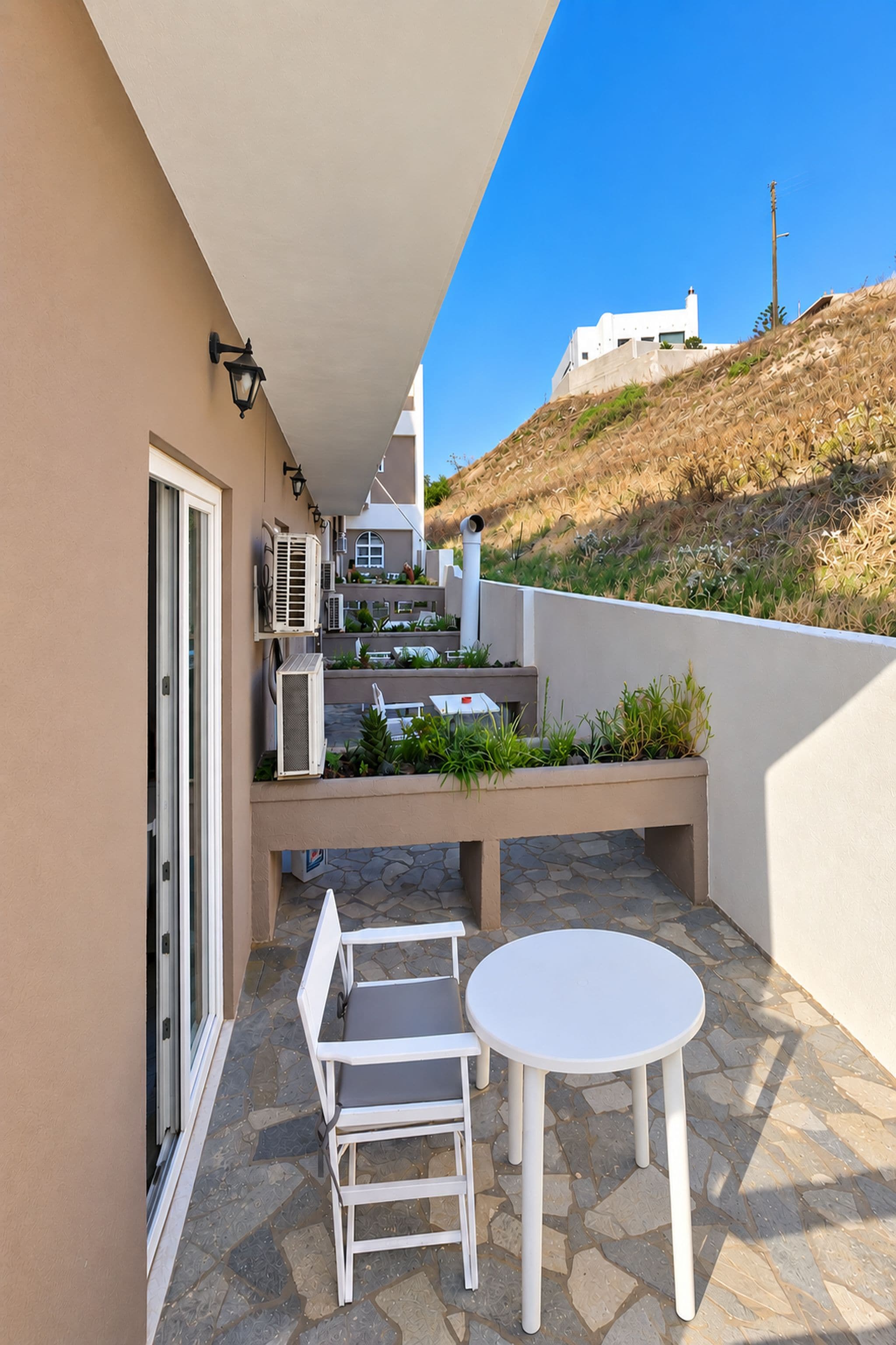 Ein Balkon mit einem weißen Tisch und Stühlen, Klimaanlagen und Blick auf eine Hügelside mit Gebäuden.