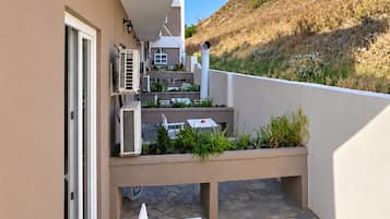 Ein Balkon mit einem weißen Tisch und Stühlen, Klimaanlagen und Blick auf eine Hügelside mit Gebäuden.