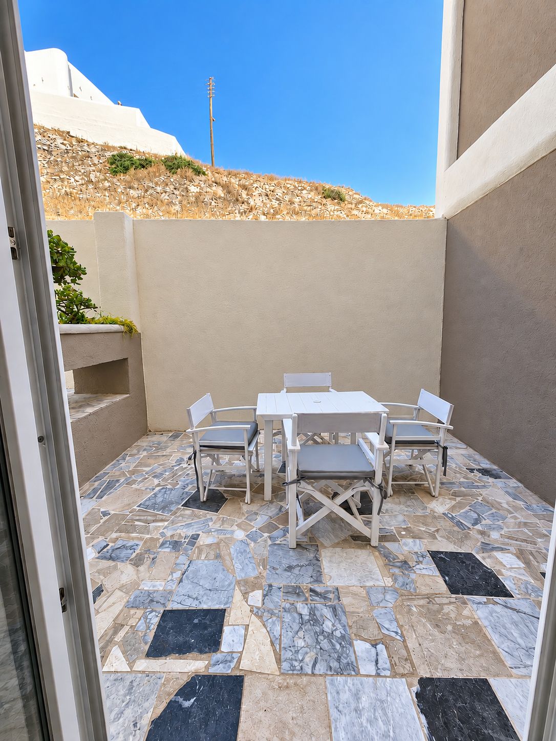 Un patio exterior con una mesa y sillas blancas, un suelo estampado y vistas a una colina.