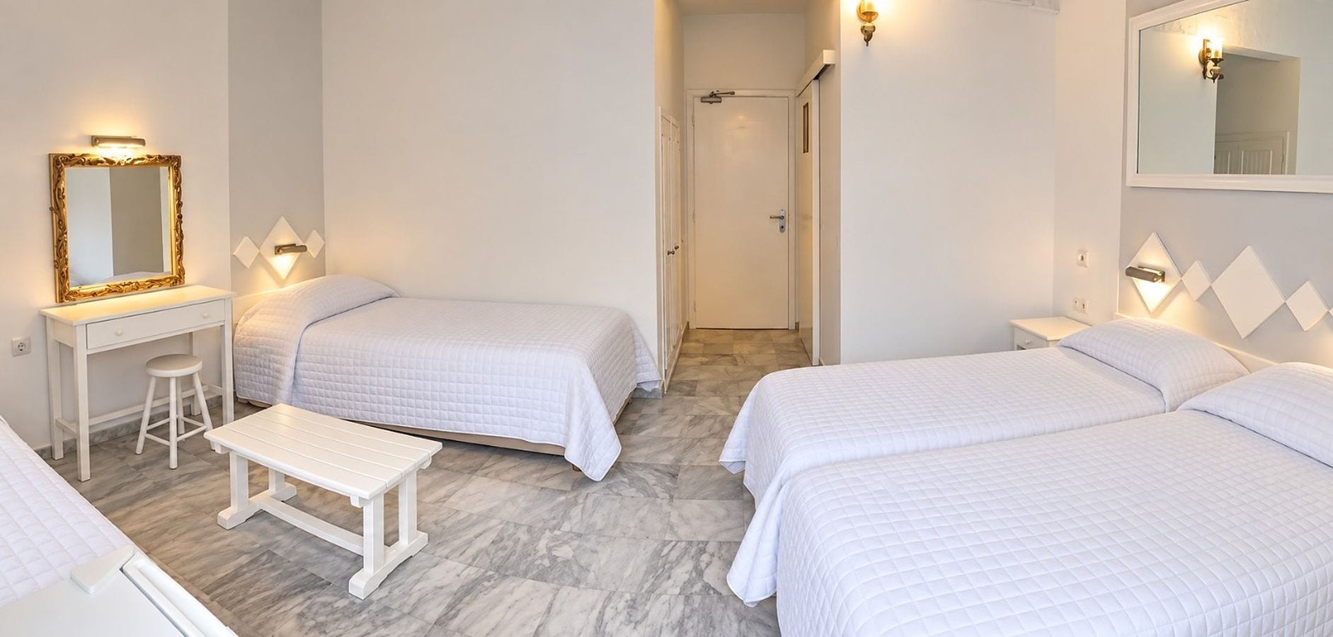 A hotel room with two beds, a desk, a bench, and a mirror.