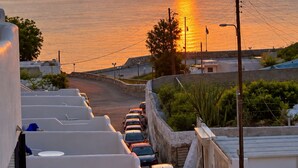 A coastal sunset view with a row of white buildings and parked cars along a street. - Porto Castello (Santorini)