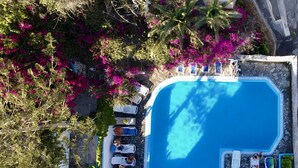 An aerial view of a swimming pool surrounded by lounge chairs and pink bougainvillea flowers. - Porto Castello (Santorini)