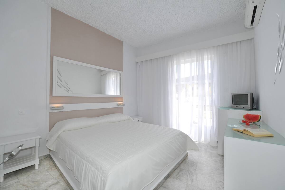 A modern hotel room with a large bed, a bedside table, a TV, and a small desk with a watermelon on it.