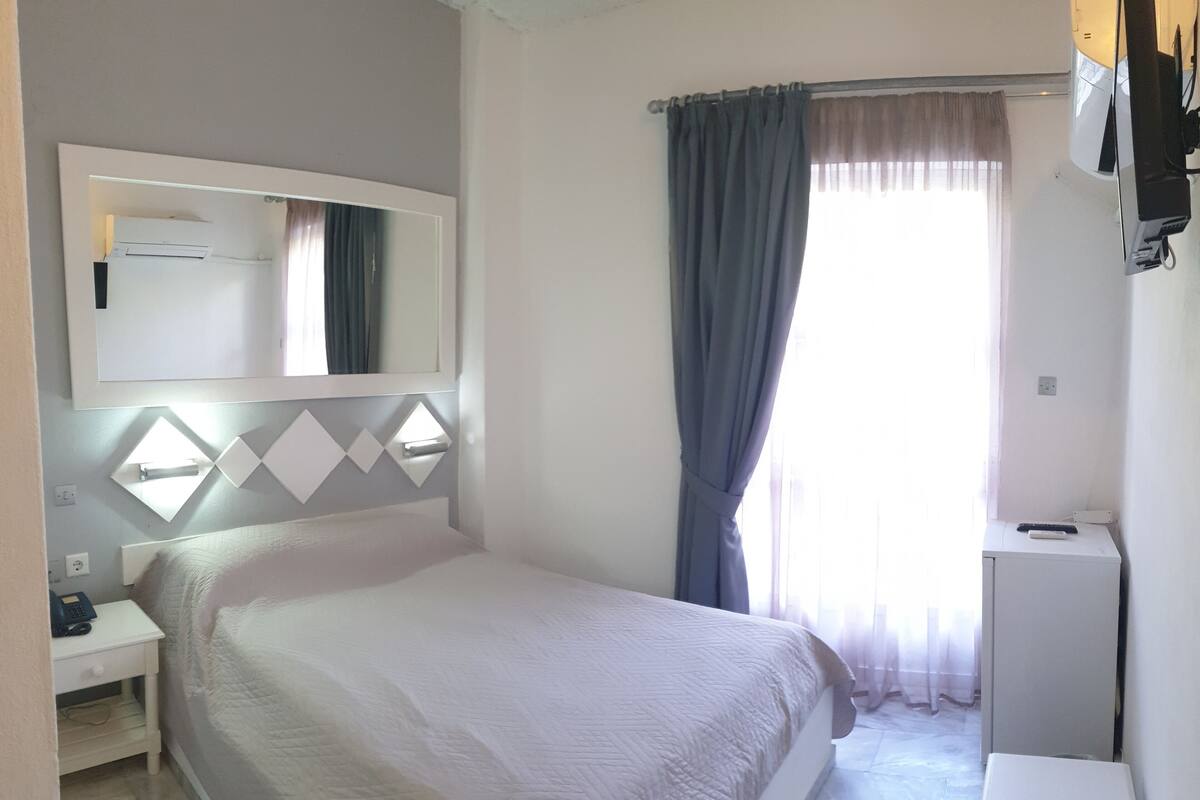 A hotel room with a bed, a bedside table with a lamp, a mirror, a desk with a computer, a TV, and a window with curtains.