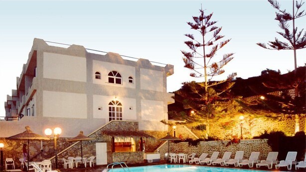A swimming pool with lounge chairs and a building with a stone facade and arched windows. - Porto Castello (Santorini)