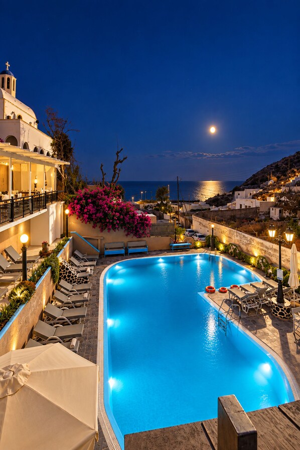 A swimming pool with lounge chairs and a view of the sea at night. - Porto Castello (Santorini)