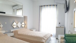 A hotel room with two beds, a small bedside table, a wall-mounted TV, an air conditioning unit, and a window with curtains. - Porto Castello (Santorini)