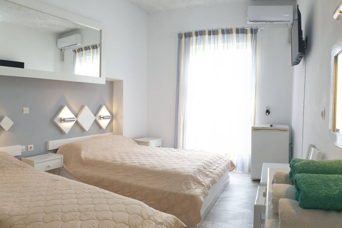 A hotel room with two beds, a small bedside table, a wall-mounted TV, an air conditioning unit, and a window with curtains.