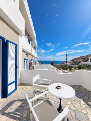 Um edifício branco com detalhes em azul, um pátio de pedra, uma mesa e cadeira brancas e vista para o mar.