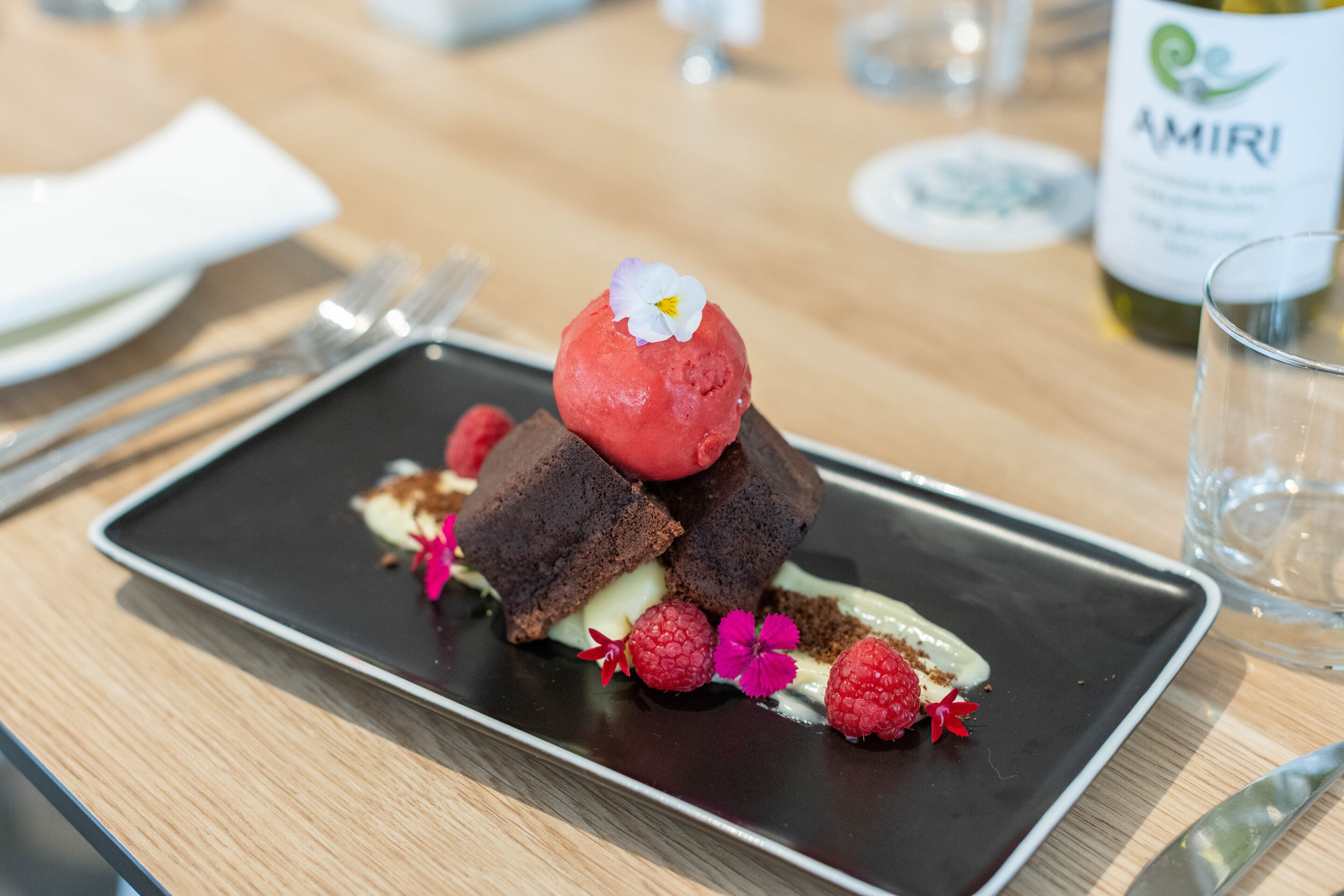 Un plato de postre con un pastel de chocolate, una bola de helado rojo, frambuesas y una flor sobre un plato rectangular.