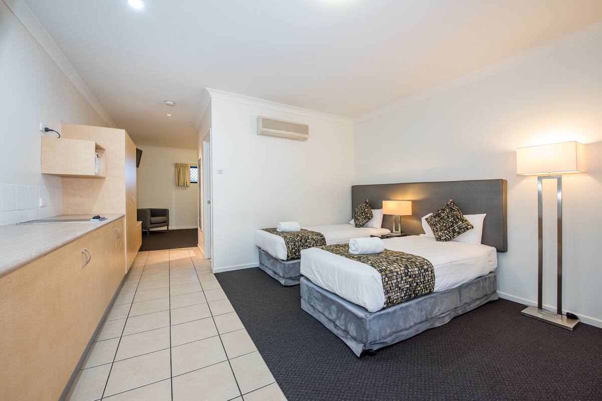 A hotel room with two beds, a desk, a chair, and a lamp.