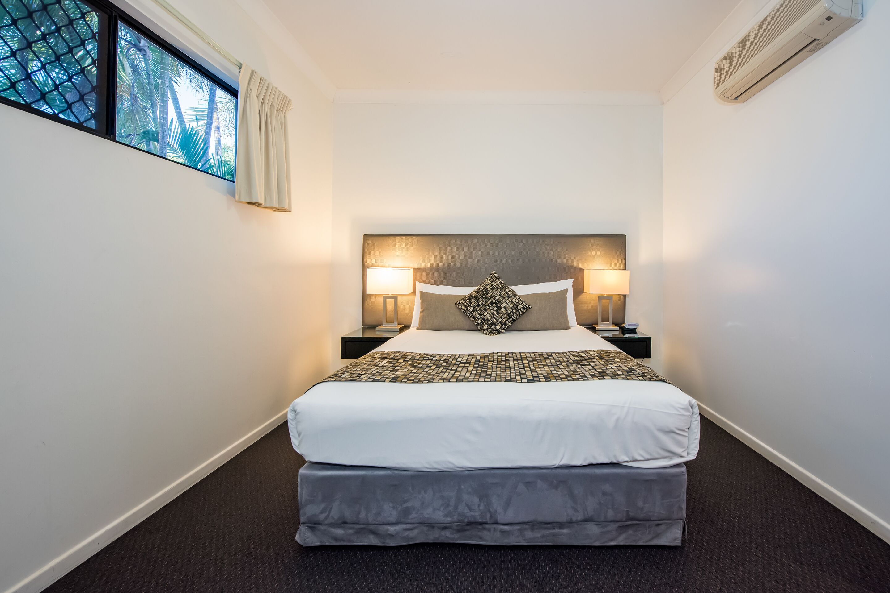 A hotel room with a large bed, two bedside lamps, a window with curtains, and an air conditioning unit.