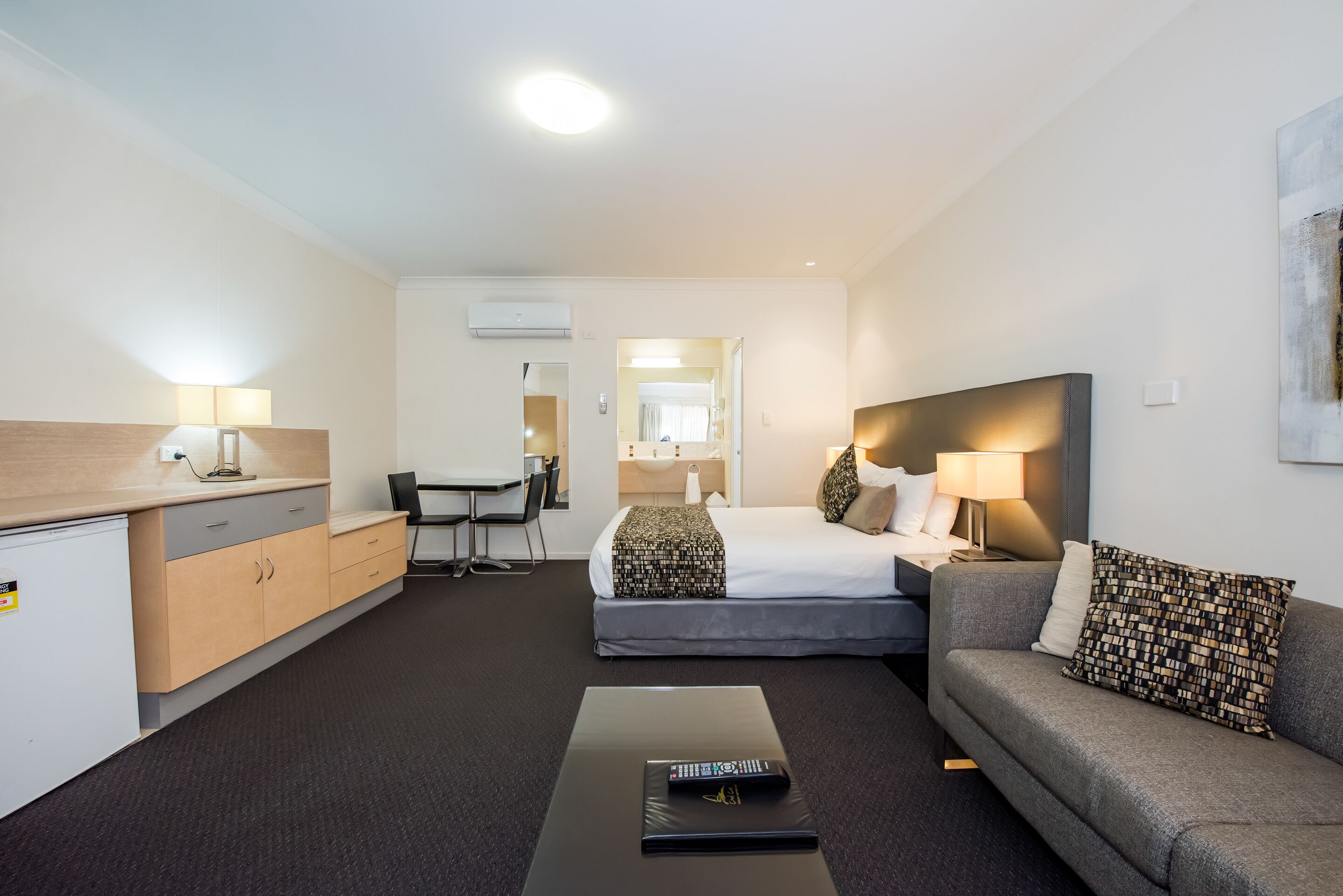 A modern hotel room with a bed, a sofa, a desk, and a small kitchenette.