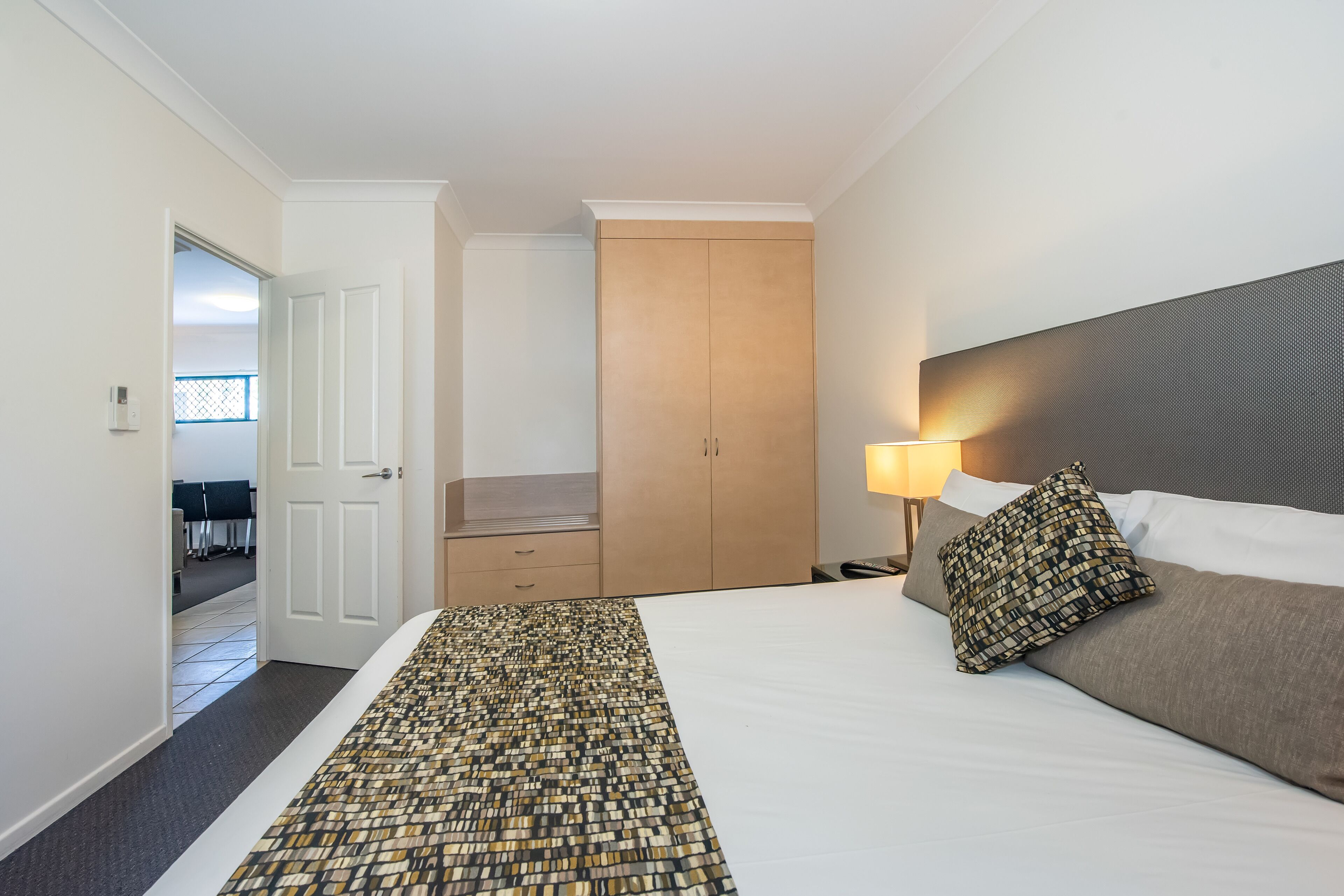 A hotel room with a bed, a nightstand, a lamp, a wardrobe, and a view of the hallway.