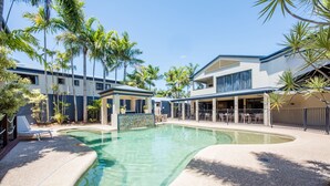 Une piscine à bord incurvé, entourée de palmiers et d’un bâtiment doté d’un patio couvert.