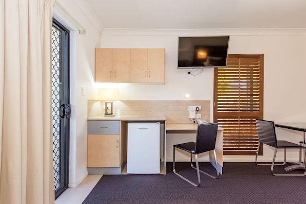A compact kitchenette with a refrigerator, a small sink, and a countertop. There is a TV mounted on the wall above the countertop and a window with blinds to the right. - Coral Cay Resort (West Mackay)
